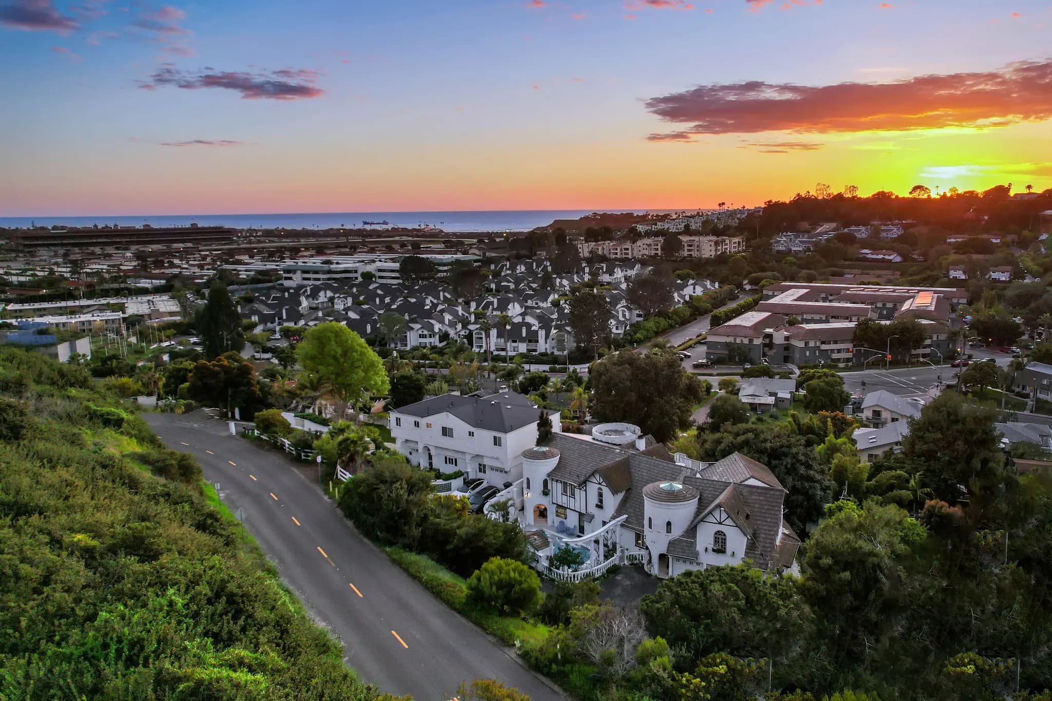 Aerial view of 915 Highland Drive at sunset, overlooking a coastal neighborhood with homes and greenery leading to the ocean.