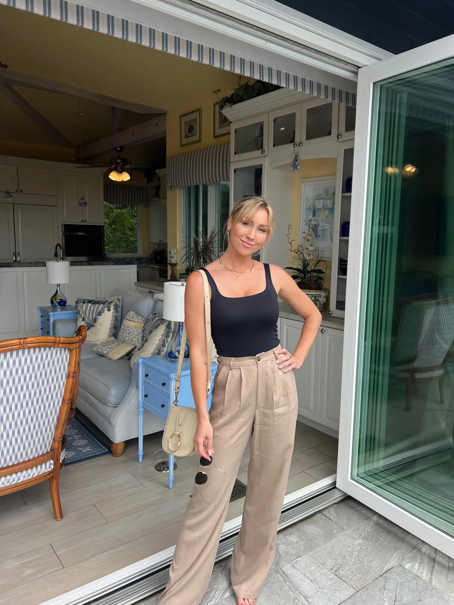 Brittany Etherton standing in front of an open glass door, wearing a black tank top and beige wide-leg pants, holding sunglasses and a beige handbag