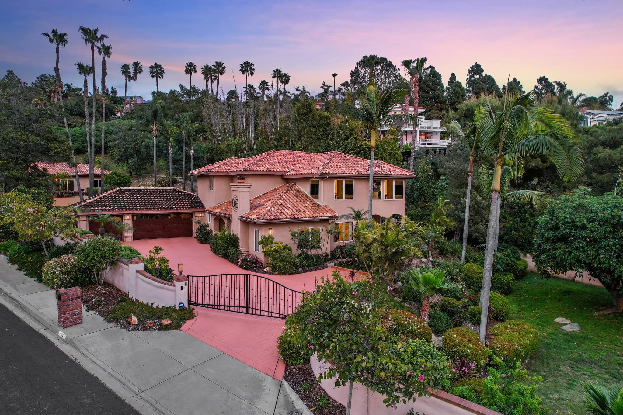 Exterior view of Cadencia Property, a Mediterranean-style home with red-tiled roofs and a gated driveway, surrounded by lush greenery and tall palm trees.