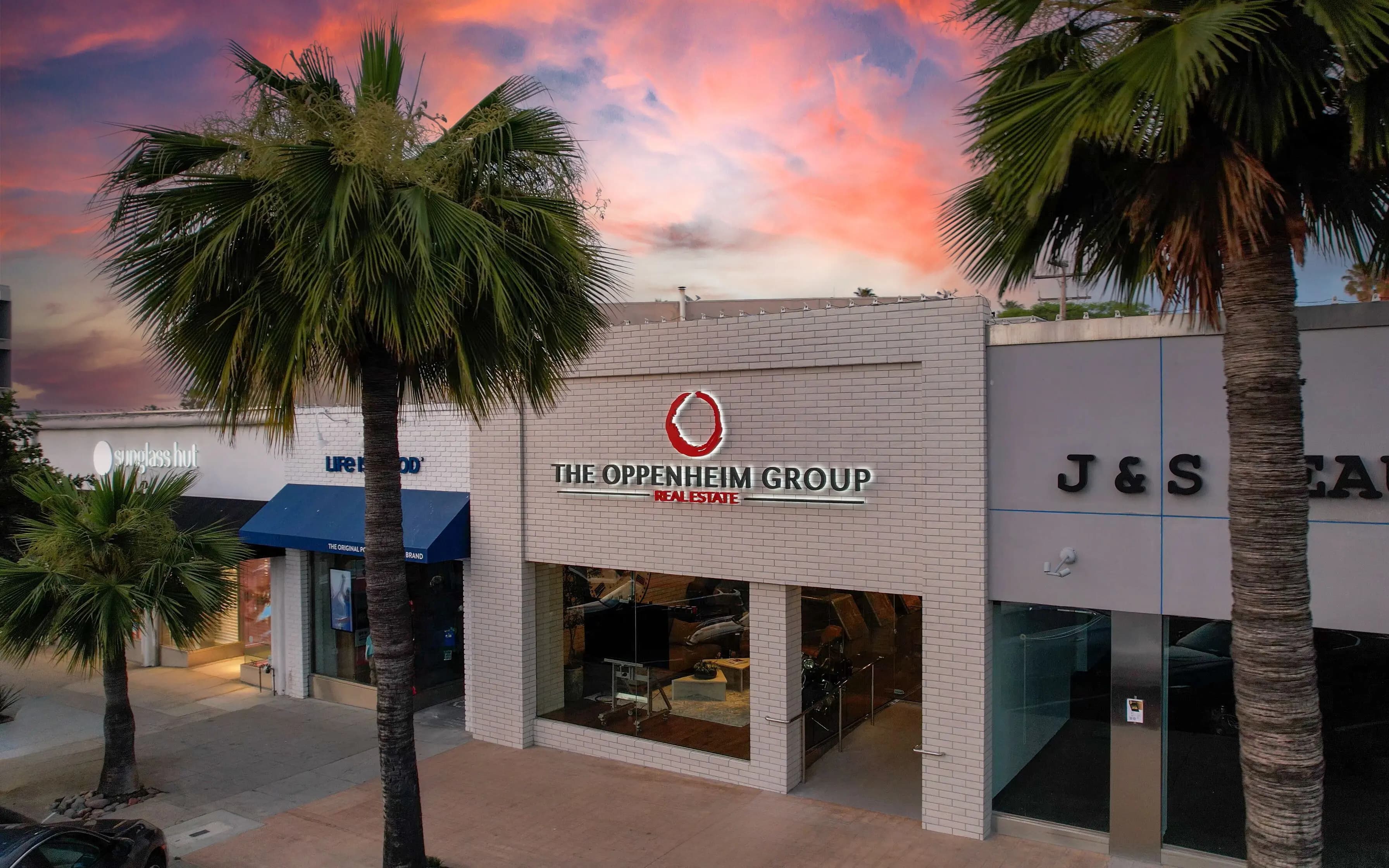 Exterior view of The Oppenheim Group Real Estate office at sunset, featuring a white brick building with the company’s logo prominently displayed.